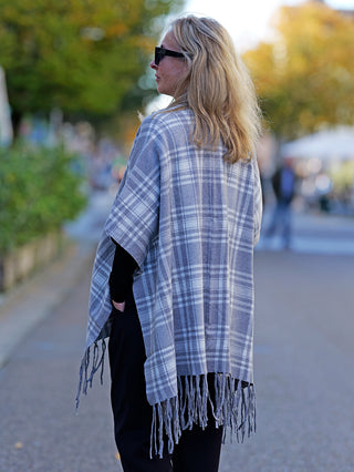 Eine Frau trägt den OFF#DLY Poncho Dalton grau, ein modisches Plaid-Poncho, perfekt für Street Fashion, mit lässigem Stil und als vielseitiges Modeaccessoire geeignet.