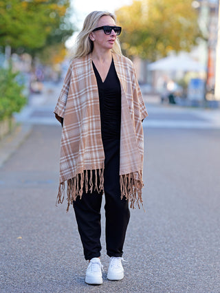 Frau trägt den OFF#DLY Poncho Dalton beige auf dem Gehweg, kombiniert mit braun-weißem Schal und weißen Sneakern. Der Poncho ist stilvoll und bequem, ideal für lässige Straßenmode.