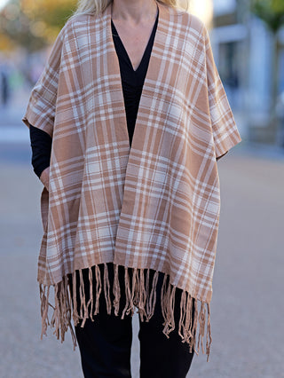 Person trägt den OFF#DLY Poncho Dalton beige mit braun-weißem Karo-Muster und Fransen, zeigt lässigen Stil. Der Poncho hat eine legere Passform und ist ideal für den Alltag.