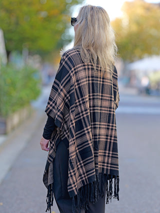 Eine Frau trägt den OFF#DLY Poncho Dalton in Schwarz/Beige, ein kariertes Modell mit lässigem Schnitt, kombiniert mit Sonnenbrille und langen, blonden Haaren, passend für Street Fashion.