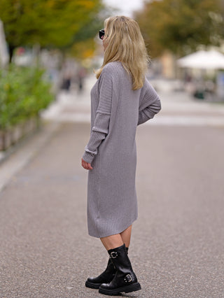Frau trägt das OFF#DLY Dress Clara grey, ein langes Strickkleid mit Rundhals, kombiniert mit schwarzen Stiefeln. Ideal für Straßenmode und lässige Anlässe.