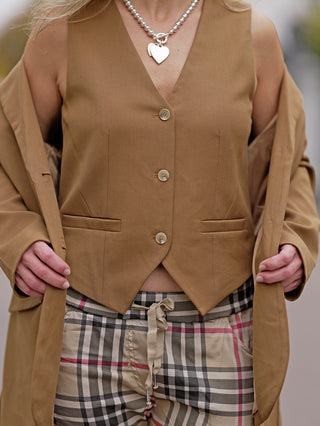 Frau trägt OFF#DLY Weste Julia camel mit seitlichen Taschen und Knopfleiste, kombiniert mit karierten Hosen. Nahaufnahme zeigt ein Herzanhänger und eine Hand.