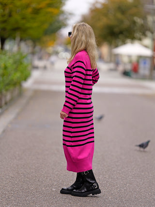 Frau trägt das OFF#DLY Ringelkleid pink mit schwarzen Streifen, kombiniert mit schwarzen Stiefeln. Das Kleid hat einen Rundhalsausschnitt und bietet eine stilvolle, lässige Passform.