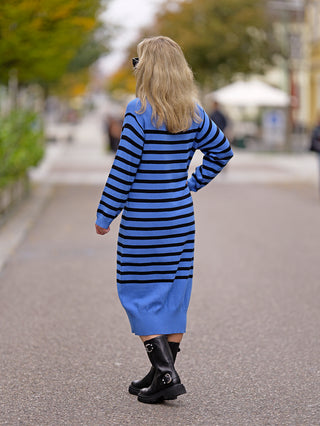 Frau trägt das OFF#DLY Ringelkleid Blue beim Spaziergang, kombiniert mit schwarzen Stiefeln. Das Kleid hat einen Rundhalsausschnitt und betont den Street-Fashion-Look. Material: 88% Viskose, 12% Nylon.