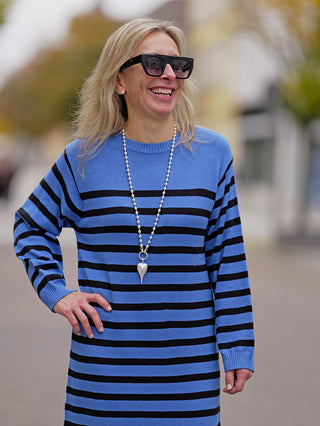 Frau trägt das OFF#DLY Ringelkleid in Blau mit schwarzen Streifen, kombiniert mit Sonnenbrille und Perlenkette. Elegantes, streifenförmiges Design ohne Taschen, ideal für lässige Anlässe. Material: Viskose und Nylon.