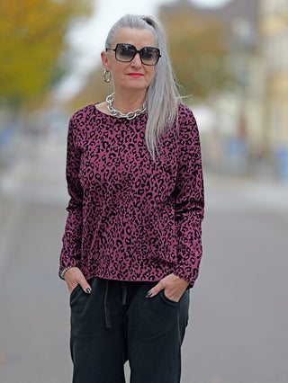Frau mit Sonnenbrille trägt das OFF#DLY Shirt Cindy burgundy, ein langärmliges Baumwollshirt mit Leo-Flockprint. Kombiniert mit schwarzer Hose, Hände in den Taschen, zeigt es Street-Fashion-Stil.