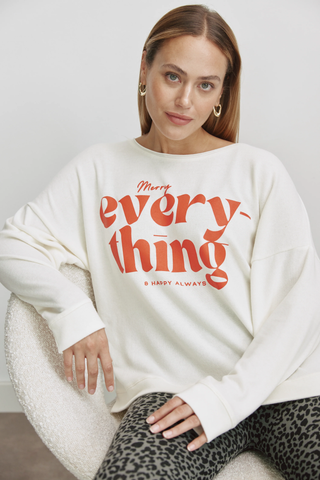 Frau sitzt im Cotton Candy Irma Tee offwhite, ein lässiges Langarmshirt mit rotem MERRY EVERYTHING Print, oversized geschnitten, ideal für vielseitige Kombinationen. Trägt goldene Ohrringe.