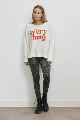 Frau trägt Cotton Candy Irma Tee offwhite, ein lässiges Langarmshirt mit rotem MERRY EVERYTHING Print, kombiniert mit Leopardenhose und schwarzen Stiefeln, betont oversized Schnitt und weiches Material.