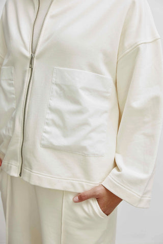 Person trägt Cotton Candy Liz Sweat, eine weiße, oversized Sweatjacke mit Troyer Kragen und aufgesetzten Webtaschen. Nahtdetails und Knöpfe sichtbar. Ideal für vielseitige Kombinationen.
