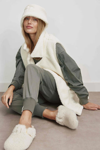 Frau sitzt auf dem Boden und trägt die Cotton Candy Lina Weste, eine oversized Teddy-Weste mit aufgesetzten Taschen und Rückenlogo, kombiniert mit weißen flauschigen Slippern.