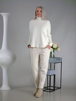 Frau trägt OFF#DLY Joggpant Doris beige mit Bundfalte und seitlichen Taschen, kombiniert mit einem weißen Pullover. Die Hose hat einen Gummibund, Kordelzug und Aufschlag am Bein.