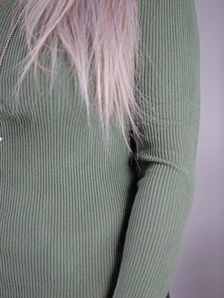 OFF#DLY Longsleeve Nina khaki in Nahaufnahme zeigt einen grünen Strickpullover mit V-Ausschnitt und geradem Schnitt, getragen von einer Frau. Sichtbar sind auch ein gestricktes Muster und eine Perlenkette.
