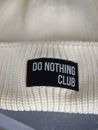 Halsüberkopf Mütze Do Nothing Club sahne, gestrickte weiße Mütze mit schwarzem Patch, aus 100% Bio-Baumwolle. Zeigt Nahaufnahme des Patches und der Strickstruktur. Einheitsgröße.