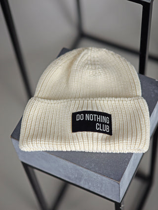 Halsüberkopf Mütze Do Nothing Club sahne: Weiße Strickmütze aus Bio-Baumwolle mit schwarzem Patch vorne, präsentiert auf einer grauen Oberfläche. Ideal für kühle Tage.