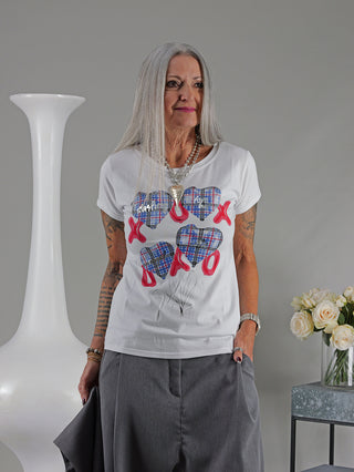 Eine Frau trägt das OFF#DLY Tee XOXO weiß/blau mit Herzmotiv. Das Shirt hat eine gerade Passform und besteht aus Baumwolle und Elastan. Im Hintergrund eine Vase mit weißen Rosen.