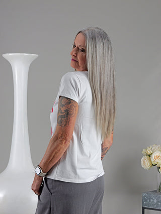 Frau im OFF#DLY Tee XOXO weiß/blau mit geradem Schnitt und Tattoos am Arm, steht neben einer weißen Vase mit rotem Deckel. Model trägt Größe S/M.
