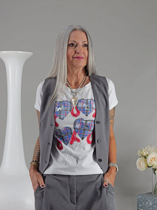 Eine Frau trägt das OFF#DLY Tee XOXO weiß/blau mit Herzmotiv, kombiniert mit grauer Weste und weißem Hemd, steht vor einer Vase mit weißen Rosen.