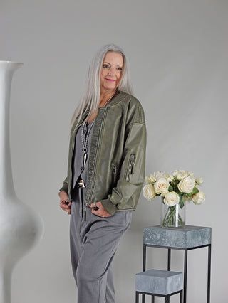 Eine Frau trägt die OFF#DLY Fake Lederjacke Sina khaki im Piloten-Stil mit Strickbund und Zippern, stehend neben einer Vase mit weißen Rosen.
