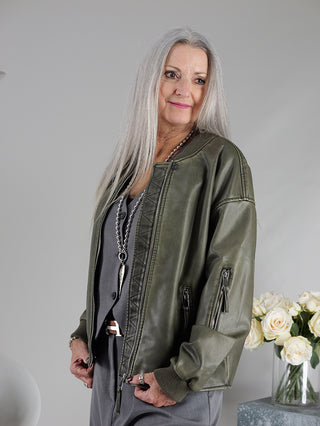 Frau mit grauen Haaren trägt die OFF#DLY Fake Lederjacke Sina khaki im Pilotenstil, mit Strickbund und Zippern, vor einer Vase mit weißen Rosen.