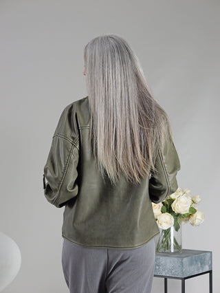 Frau mit grauen Haaren trägt die OFF#DLY Fake Lederjacke Sina khaki, eine sportive Pilotenjacke mit Strickbund und Zippern, während sie eine Vase mit weißen Rosen berührt.