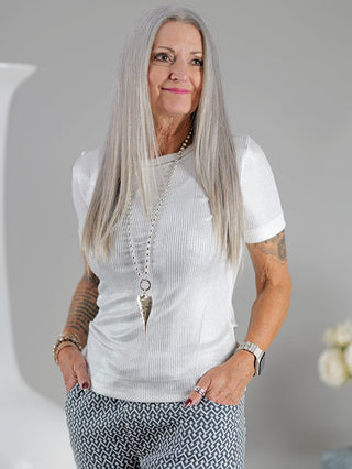 Frau mit langem grauen Haar trägt das OFF#DLY Top Sindy weiß/silber mit silbernem Lurex-Faden und Rundhalsausschnitt, ergänzt durch eine auffällige Halskette.