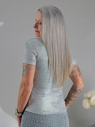 Eine Frau trägt das OFF#DLY Top Sindy grau/silber, ein stylisches Feinripp-Top mit silbernem Lurex-Faden und Rundhalsausschnitt, neben sichtbaren Tattoos auf ihrem Arm.