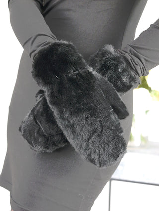 Nahaufnahme einer Person mit einem schlichten schwarzen Kleid, die oversized, flauschige schwarze OFF#DLY Handschuhe aus Fakefur trägt. Die Fäustlinge bestehen aus 100% Polyester und sind maschinenwaschbar.