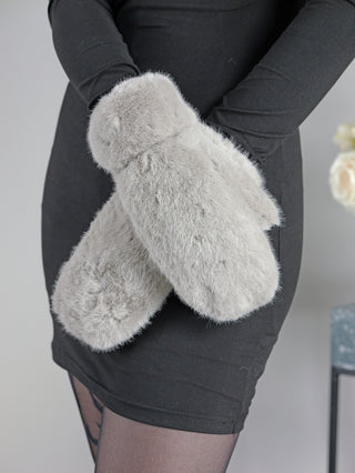 Eine Person hält die OFF#DLY Handschuhe grau, flauschige Fäustlinge aus Fakefur, in jeder Hand. Sie trägt ein schwarzes langärmliges Oberteil und durchsichtige schwarze Strumpfhosen.
