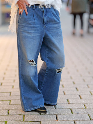 Person trägt Gitta Banko Jeans Raven Destroy mit weitem Schnitt und zerstörtem Denim-Look, sichtbare Löcher an den Knien, seitliche und hintere Taschen.