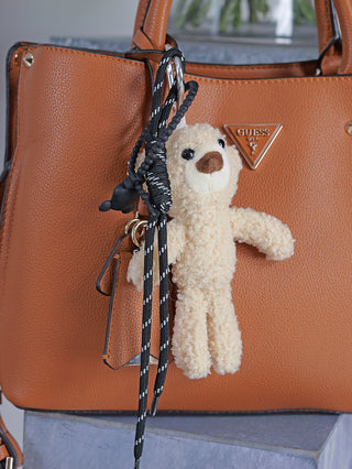 OFF#DLY Bag Charm Teddy beige: Ein cremefarbener Plüsch-Teddybär-Anhänger mit lockiger Fellstruktur, befestigt an einer schwarzen und weißen Kordel, hängt an einer Guess-Tote-Tasche aus braunem, genarbtem Leder.