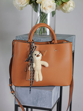 OFF#DLY Bag Charm Teddy beige: Ein strukturierter, cognacbrauner Leder-Shopper von GUESS mit einer cremefarbenen Teddy-Anhänger an einem Griff, ergänzt durch zwei schwarze und weiße Schnüre.