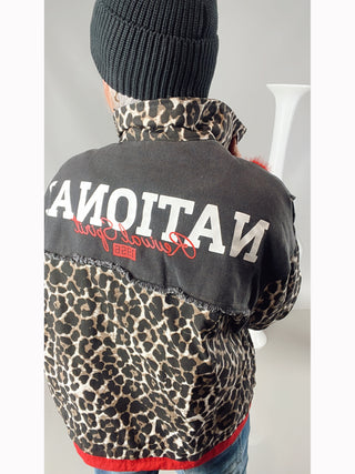 Person trägt die OFF#DLY National Jacke black/leo mit Leopardenmuster, rotem Bund, aufgesetzten Taschen und Rückenprint NATIONAL. Oversized-Stil mit verdeckter Knopfleiste und Zipper.