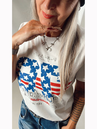 Frau trägt OFF#DLY Tee Vintage weiß mit blau/rot/silbernem Print im US-Stil, kombiniert mit blauer Jeans. Entspanntes, gerades Design, ideal für Größen 34-40.