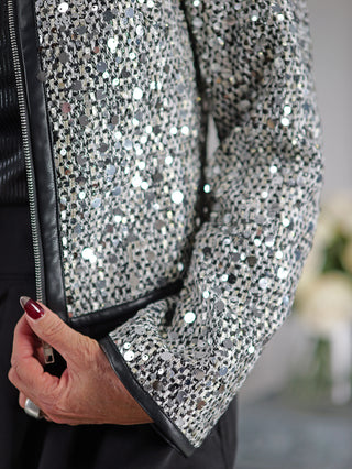 Person trägt eine OFF#DLY Jacke Mila silber/black mit Zipper und Paillettenbesatz, hält eine schwarze Tasche. Fingernägel und Ring sind sichtbar.