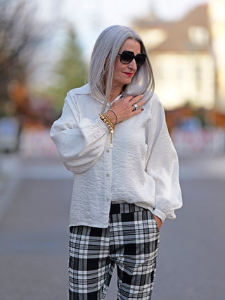 Frau mit grauem Haar trägt die OFF#DLY Bluse Jutta white mit weiten Ärmeln, kombiniert mit schwarz-weißen karierten Hosen und goldenen Armbändern.