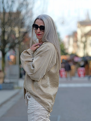 Frau mit langen grauen Haaren trägt die OFF#DLY Bluse Jutta beige, glänzende Optik, weite Ärmel, Knopfleiste, leicht oversized geschnitten. Sie kombiniert die Bluse mit Sonnenbrille und Goldschmuck.