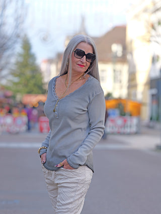 Frau auf der Straße trägt OFF#DLY LS Maribel grau Longsleeve mit V-Ausschnitt und Spitzenbesatz, sichtbar sind Sonnenbrille und Kette. Hände in Taschen, stilvoll und lässig.