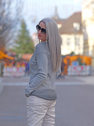 Frau mit langen grauen Haaren und Sonnenbrille trägt den OFF#DLY LS Maribel grau Longsleeve mit V-Ausschnitt und Spitzenbesatz, während sie lässig in einer urbanen Umgebung steht.