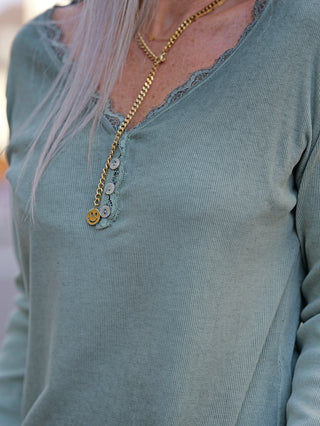 Frau trägt die OFF#DLY LS Maribel matcha Halskette mit Smiley-Anhänger, sichtbar über einem V-Ausschnitt mit Spitzenbesatz und angedeuteter Knopfleiste. Fokus auf das modische Accessoire.