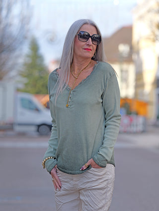 Frau im Freien trägt das OFF#DLY LS Maribel matcha Longsleeve mit V-Ausschnitt, Spitzenbesatz und angedeuteter Knopfleiste, Hände in Taschen, Sonnenbrille als Modeaccessoire.