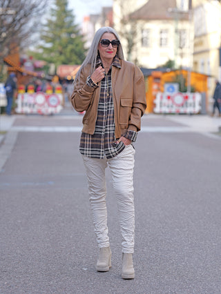 Frau mit grauen Haaren trägt die OFF#DLY Fake Leather Jacket camel, eine tanfarbene Lederjacke im Pilotenstil mit Strickbund und aufgesetzten Taschen, kombiniert mit modischen Accessoires in einer belebten Fußgängerzone.