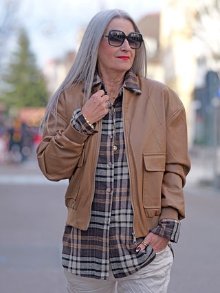 Frau trägt eine camelbraune OFF#DLY Fake Leather Jacket: Bomberstil, Reißverschluss, Klappentaschen, kombiniert mit einem langen karierten Hemd und hellen Joggern. Accessoires: schwarze Sonnenbrille, goldener Schmuck.