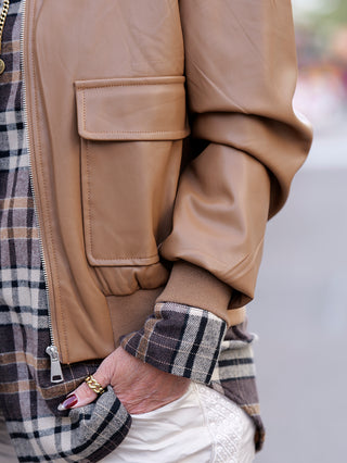 Nahaufnahme einer Frau, die die OFF#DLY Fake Leather Jacket camel trägt, mit goldfarbenem Ring an der Hand. Die Pilotenjacke hat einen Strickbund und aufgesetzte Taschen.