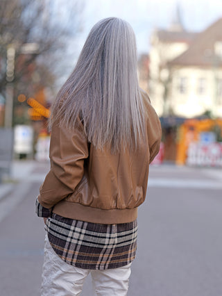 Frau mit langen grauen Haaren trägt eine OFF#DLY Fake Leather Jacket camel im Pilotenstil mit Strickbund und aufgesetzten Taschen, fotografiert von hinten.
