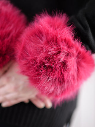 Nahaufnahme von zwei flauschigen, pink-roten Kunstfell-Pompons am OFF#DLY Pulswärmer pink. Teil eines schwarzen Stoffes ist sichtbar, möglicherweise ein Ärmel oder eine Tasche, Hintergrund unscharf.