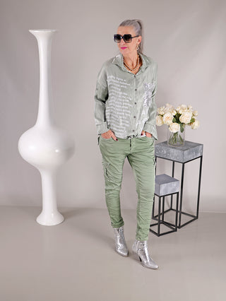 Frau trägt die OFF#DLY Bluse Ulrike matcha mit langen Ärmeln und Pailletten-Brusttasche, stehend vor einer Vase mit weißen Rosen.