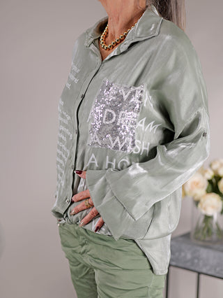 Frau trägt die OFF#DLY Bluse Ulrike matcha, eine glänzende Longbluse mit Print, Pailletten-Brusttasche und Knopfleiste. Modell mit geradem Schnitt aus 100% Viskose.