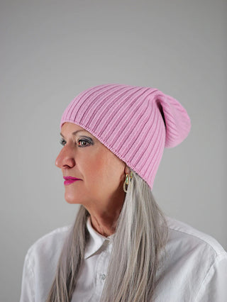 Ältere Frau trägt die OFF#DLY Mütze Anna rosa, eine gerippte, hellrosa Strickmütze. Sie kombiniert sie mit goldenen Creolen, dunklem Eyeliner und pinkem Lippenstift, passend zu ihrem weißen Hemd.