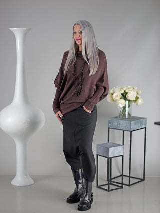 Eine Frau steht neben einer Vase; sie trägt die OFF#DLY Pant Susana black, ein klassischer Rock aus Denim mit Nieten vorne, kombiniert mit einem langen schwarzen Rock und Stiefeln.