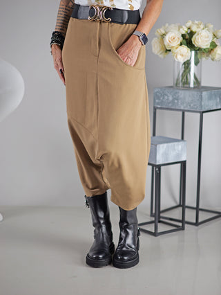 Person trägt OFF#DLY Pant Susana camel, eine lässige Haremshose mit großer Einschubtasche links, Gummibund hinten, vorne Knopf und Zipper, kombiniert mit schwarzen Stiefeln.